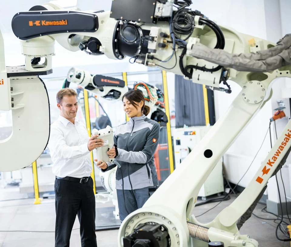 Két ember egy nagy ipari robotkar mellett áll egy világos műhelyben.