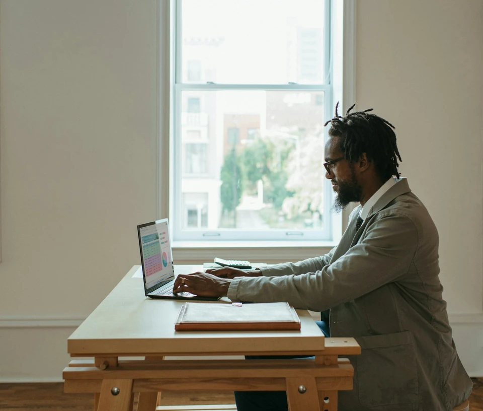 A man at a wooden desk by a window filling in a spreadsheet on his laptop, a calculator and a folder beside him.